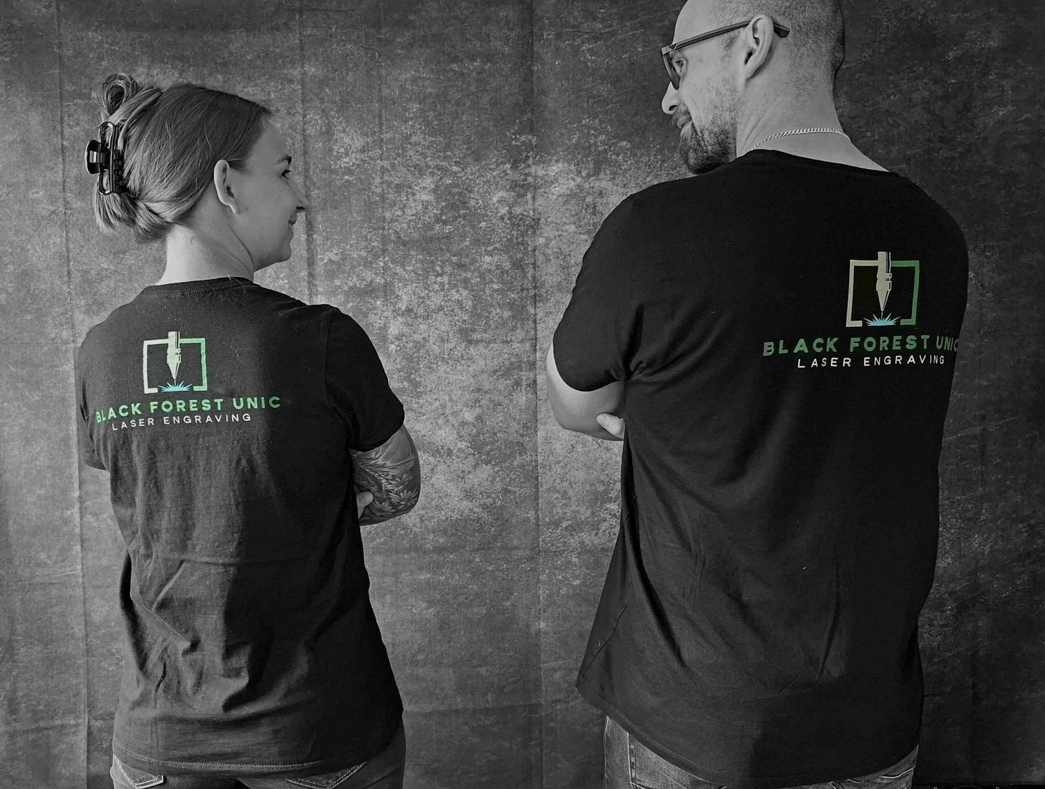 Das Bild zeigt zwei Personen von hinten, die schwarze T-Shirts mit dem Logo „BLACK FOREST UNIC – LASER ENGRAVING“ tragen. Das leuchtend grüne Firmenlogo und der Schriftzug bilden einen starken Kontrast zum dunklen Stoff. Die Pose wirkt authentisch und kollegial. Das Foto symbolisiert Zusammenhalt und Markenidentität im Lasergravur-Bereich.