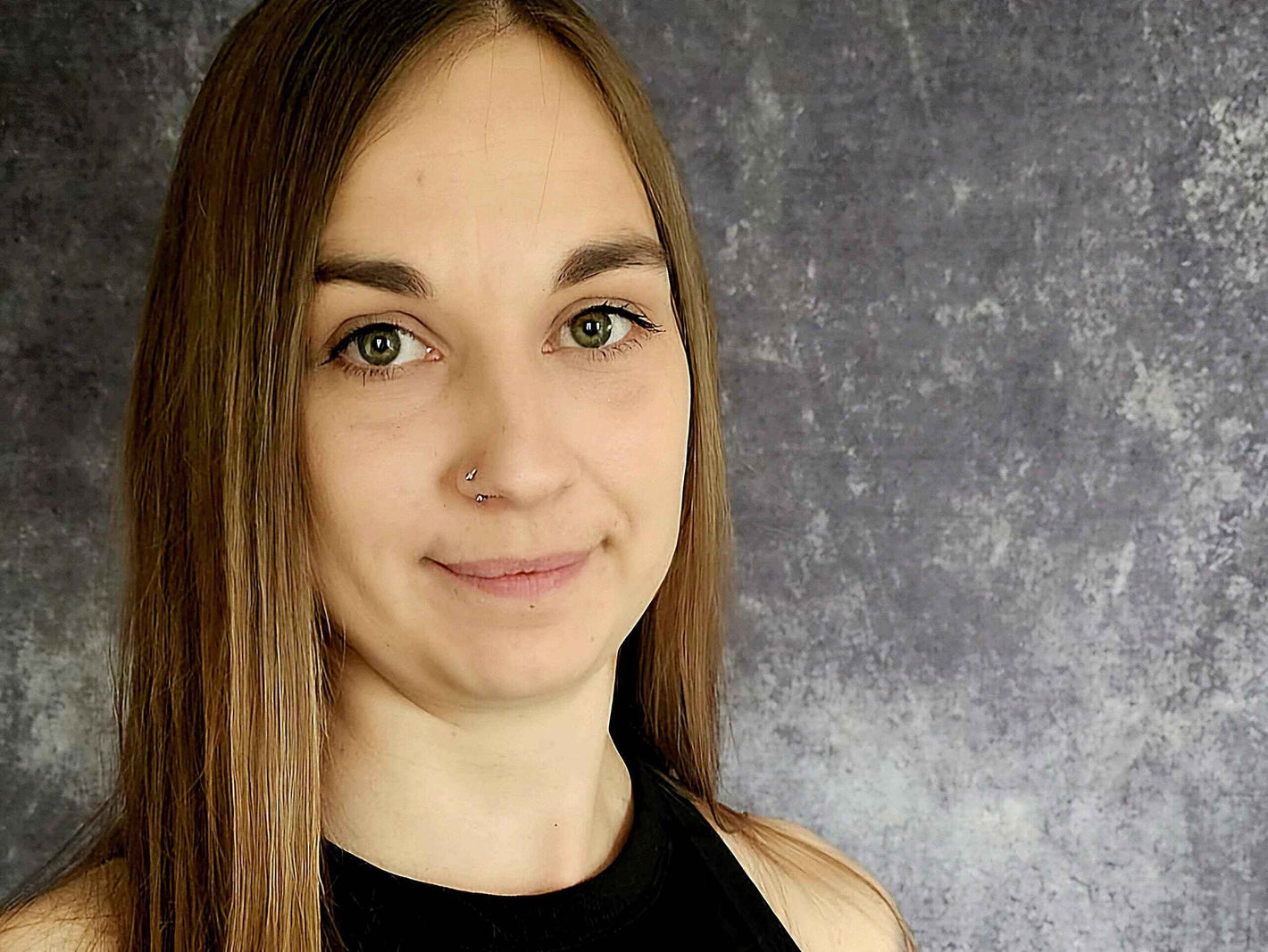 Ein professionelles Porträt einer Frau mit glatten, braunen Haaren und einem Nasenpiercing, aufgenommen vor einem neutralen grauen Hintergrund. Sie trägt ein schwarzes Oberteil und blickt ruhig in die Kamera. Das Bild vermittelt Offenheit und Persönlichkeit.