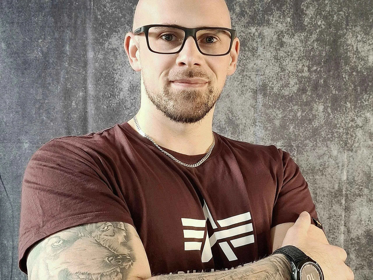 Ein professionelles Porträt eines Mannes mit Glatze, Brille und Tattoos auf den Armen, aufgenommen vor einem neutralen grauen Hintergrund. Er trägt ein T-Shirt mit Logo und eine Uhr am Handgelenk. Seine Körperhaltung mit verschränkten Armen wirkt entschlossen und sympathisch zugleich.